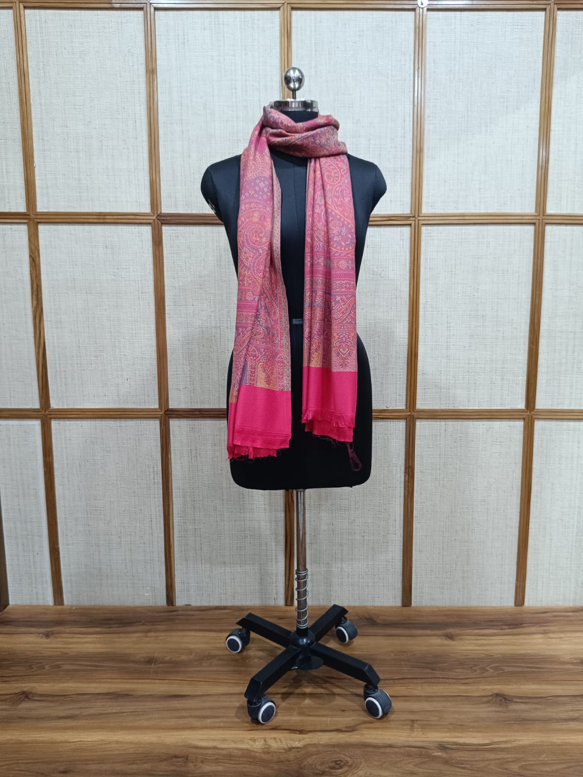 A pink and red patterned scarf with solid magenta ends is draped on a black dress form mannequin, set against a light beige paneled wall and wooden floor.